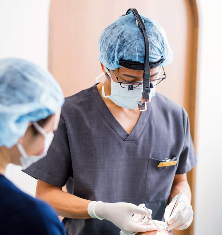 手術帽とマスクを着用した医師が、器具を手に取り施術準備を行う真剣な医療現場の様子